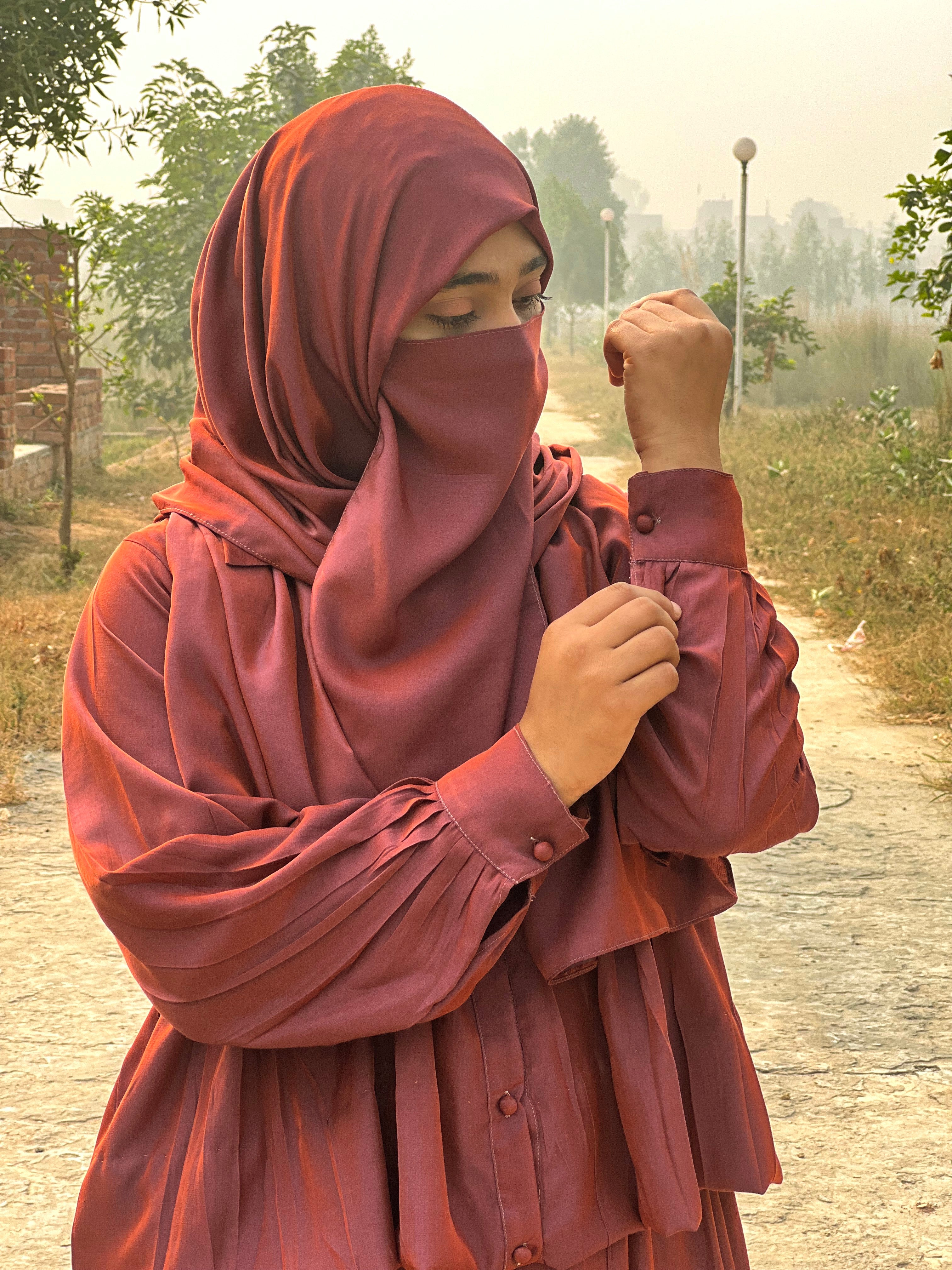 Person wearing a red hijab and dress outdoors on a path with trees and grass.