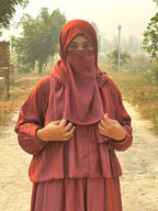 Person wearing a red hijab and dress standing outdoors on a path with trees in the background.