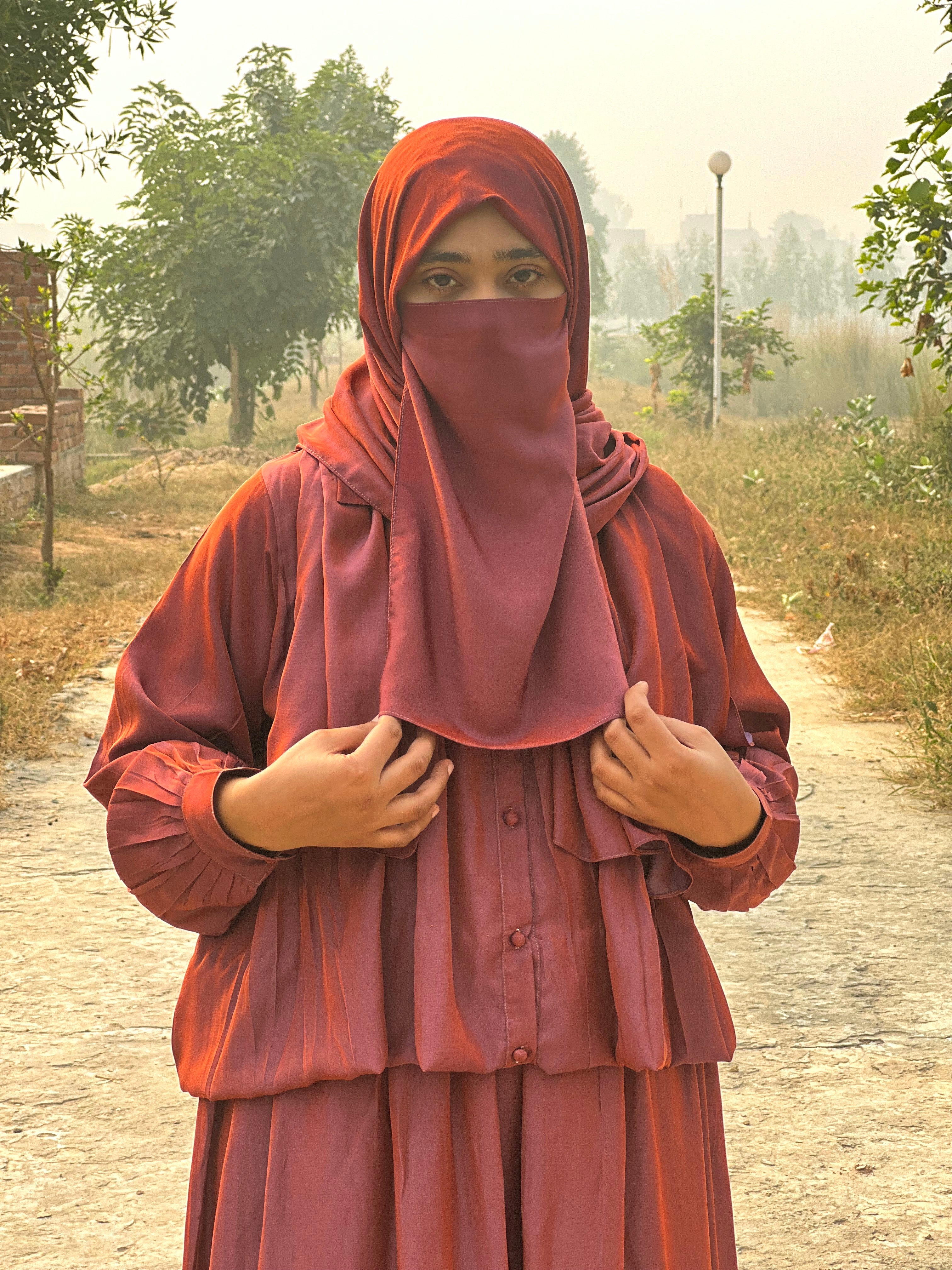 Person wearing a red hijab and dress standing outdoors on a path with trees in the background.